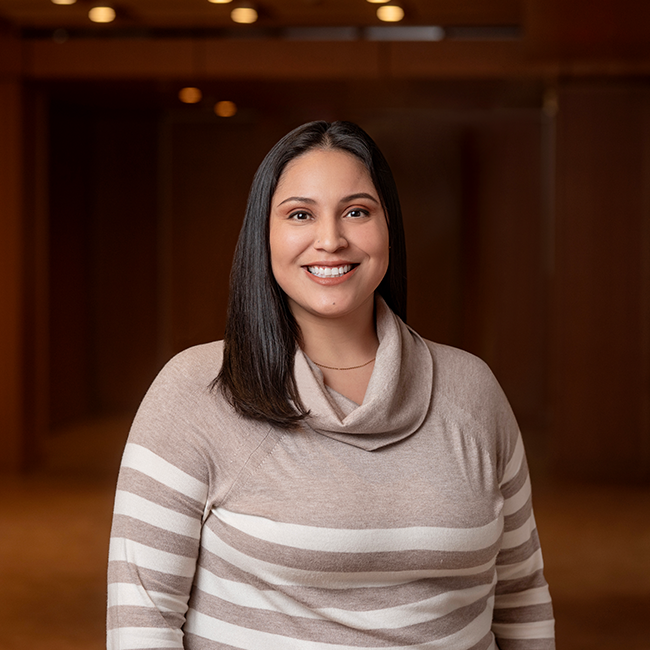 A smiling tan-skinned woman named Gaby Alvarado with shoulder-length black hair, wearing a beige and white striped sweater, and standing in a room with dark wood details.