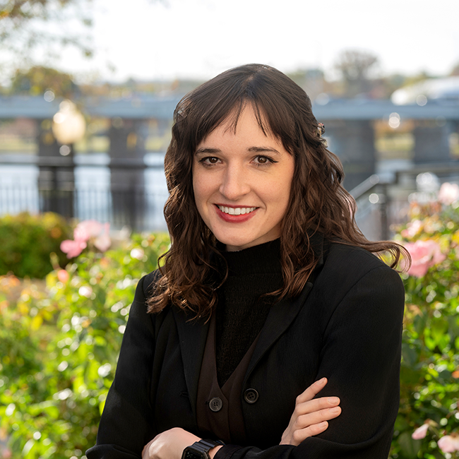 A light-skinned female with brown hair wearing a dark outfit against an outdoor background.