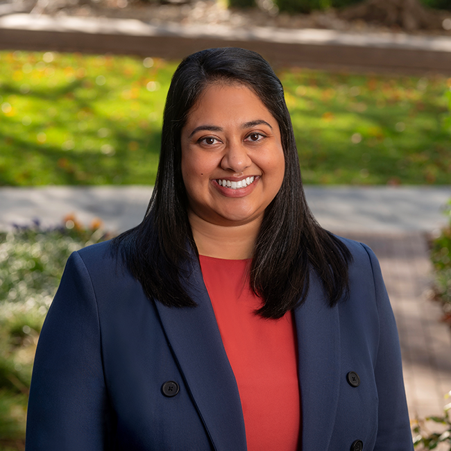 Smiling female-presenting person of color with dark hair named Ishita Das wearing a red shirt and a blue blazer against an ambiguous outdoor background.