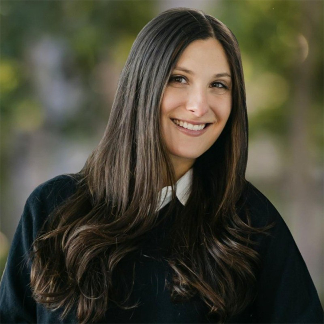 An image of a smiling adult woman named Jenny Sorin, who is  an associate director of Business and Program Development at the Milken Institute. She has long brunette hair, light white skin, and is wearing a black sweater with a white collar.