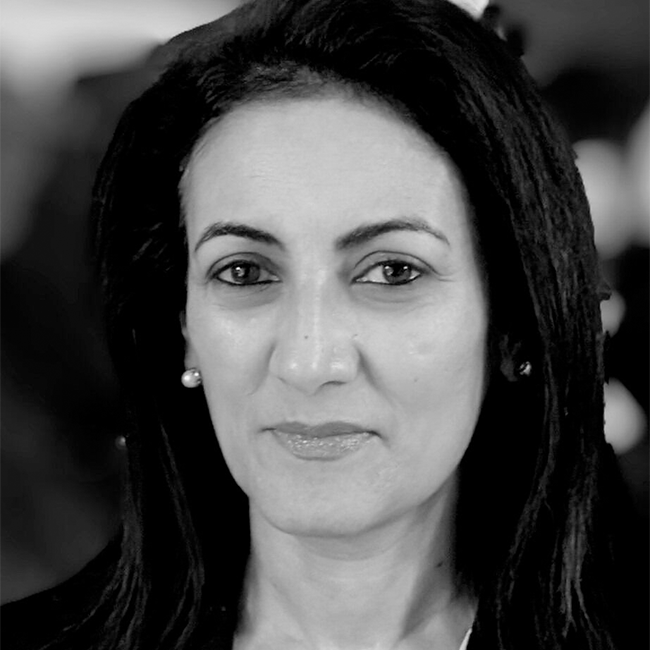 A black and white image of an adult woman with white-ish tan-ish skin and dark hair. She is wearing pearl earrings, a black shirt, and slightly smiling at the camera.