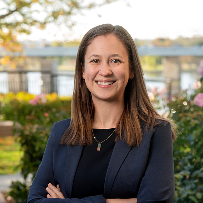 A smiling light-skinned, brunette, female-presenting individual named Kelsey Barcomb wearing a blue blazer and a black shirt against a blurred outdoor background.