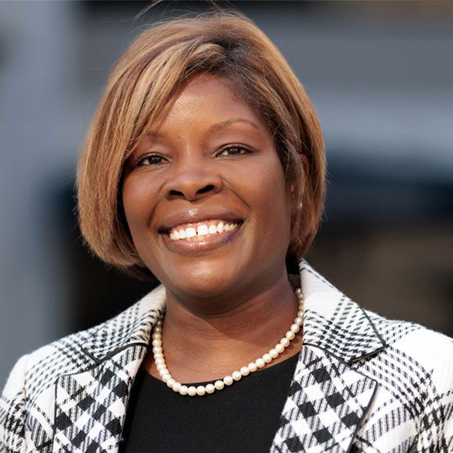 A headshot of a woman with brown skin, brown eyes, and short bobbed brunette hair. She is smiling at the camera and wearing a black and white patterned blazer and a pearl necklace with a black shirt.