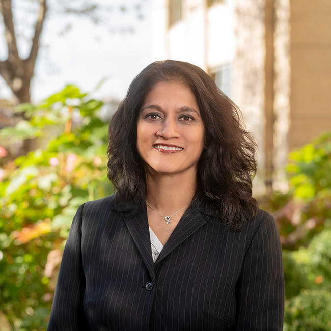 A smiling darker-skinned female named Mala Persaud with medium length black hair wearing a black blazer against an ambiguous outdoor background.