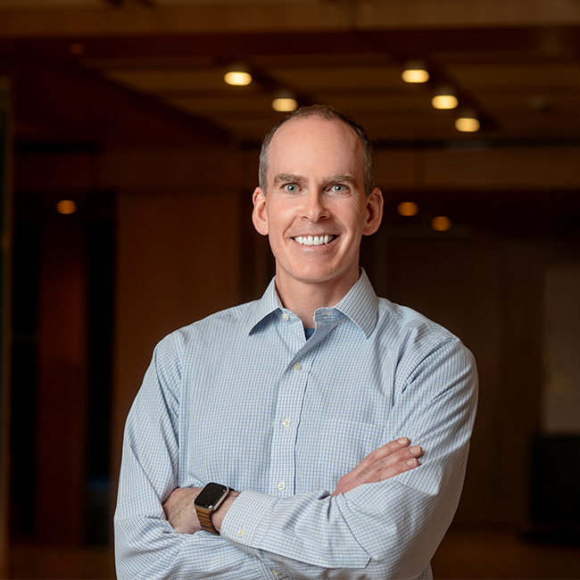 A smiling light-skinned man named Marc McDonald with a bald head, wearing a light blue button-down shirt, and standing with his arms crossed in a room with dark wood details.