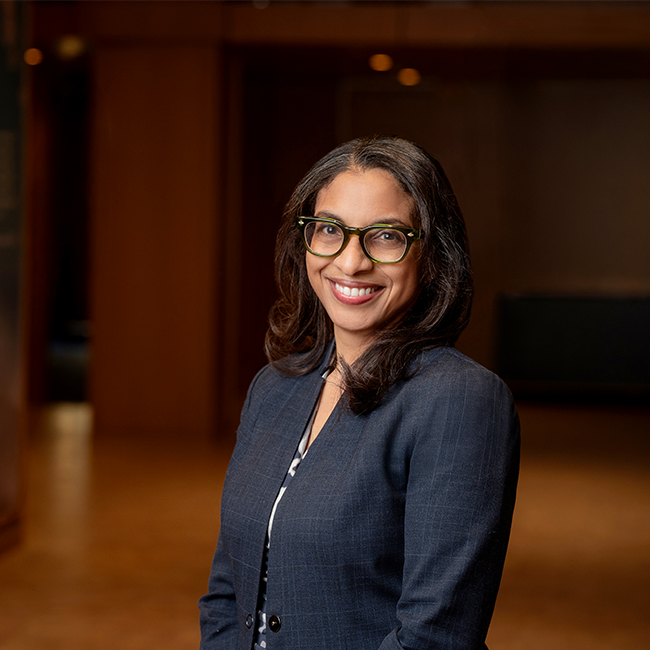 A smiling dark-skinned woman named Martine Polycarpe with long dark hair, wearing a black blazer and glasses, and standing in a room with dark wood details.