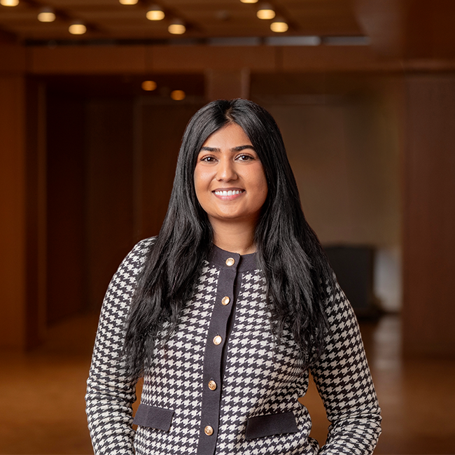 An image of a woman named Noha Shaikh who is a Senior Associate of Milken Institute Strategic Philanthropy. She is a young woman with long straight black hair, tan skin, and dark eyes. She is smiling at the camera and wearing a Houndstooth black and white patterned sweater with gold buttons, standing in a room with dark wood.