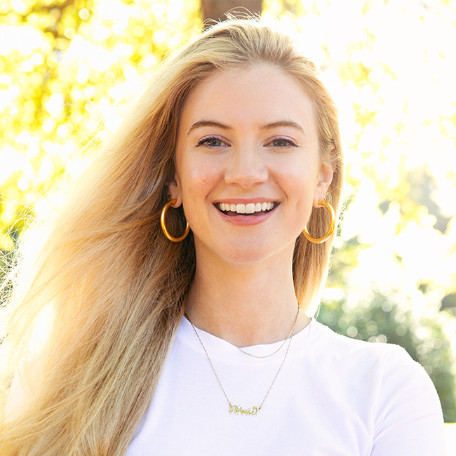 An image of a smiling adult woman named who is an associate director on the Business and Program Development team at the Milken Institute. She has long flowing blonde hair, light white skin, and she is wearing a white shirt and gold hoop earrings against a blurred outdoor background.