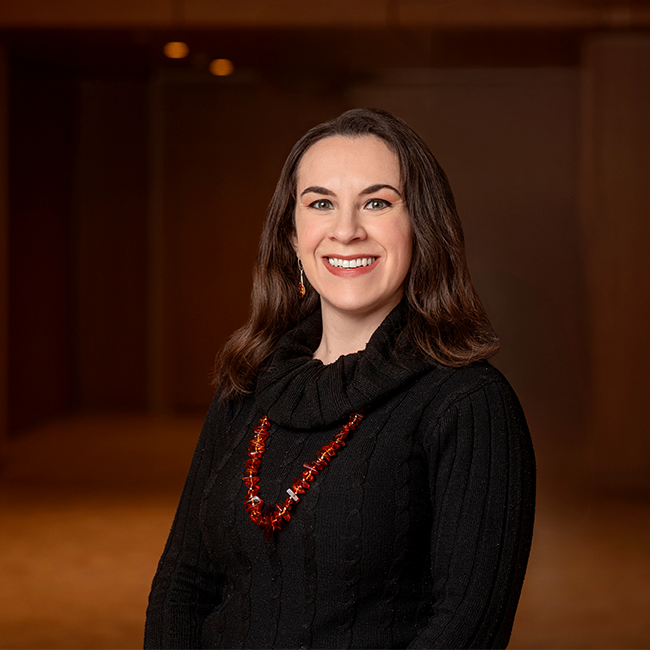 A smiling light-skinned woman named Sarah ElShafie with shoulder-length brunette hair, wearing a black sweater with a long red necklace, and standing in a room with dark wood details.