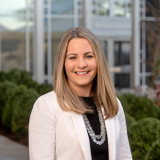 An image of a woman with white skin and shoulder-length blonde hair named Sylvie Raver. She has a white suit jacket and a black shirt and she is smiling at the camera. 