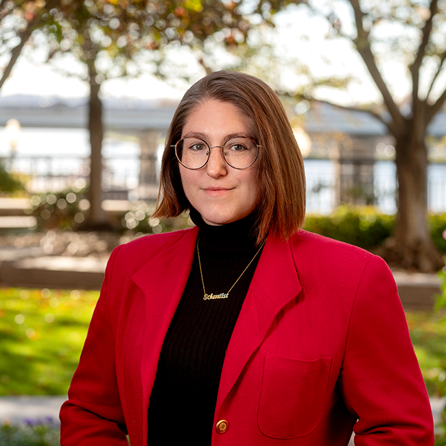 An woman named Taylor Evans white skin, bobbed auburn hair and glasses. She is wearing a red blazer with a black shirt and standing outside.