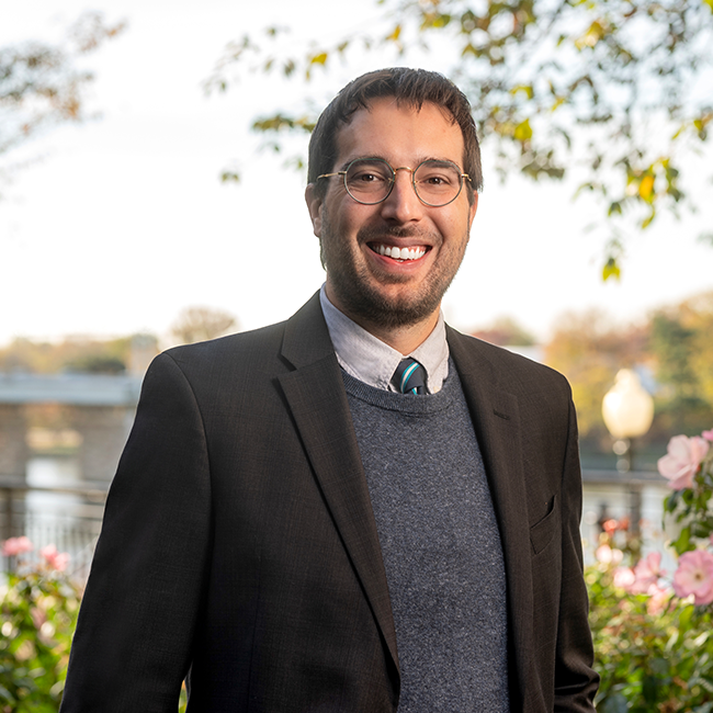 An image of an adult man named Tom Jurkiw. He has white skin, short brown hair, and a brunette beard. He is wearing glasses, a black blazer, a blue sweater underneath, and a tie, and smiling at the camera with an ambiguous outdoor background.