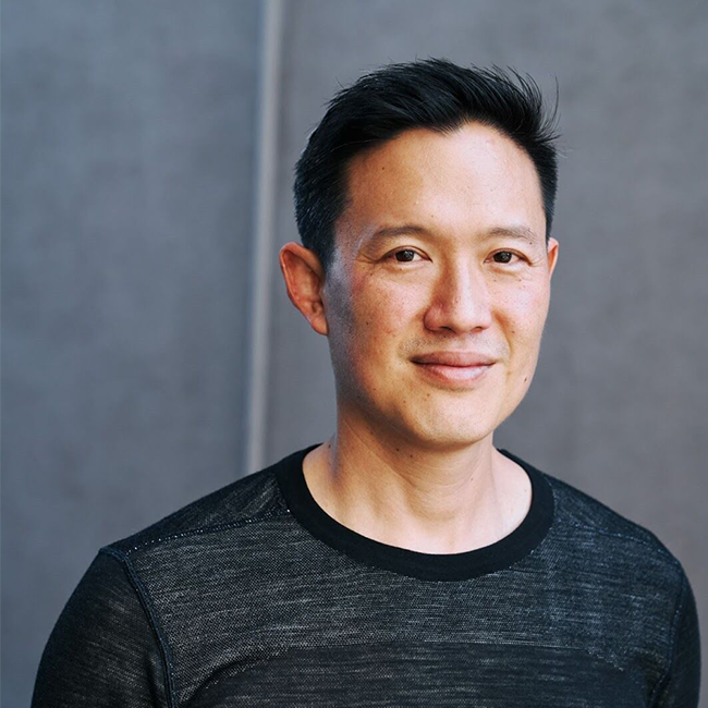 An image of a man named Avery Ching who is CEO, CTO, and Co-Founder of Aptos Labs. He has white skin, short black hair, brown eyes, and is Asian in appearance. He is wearing a black shirt, standing against a gray background and smiling at the camera.