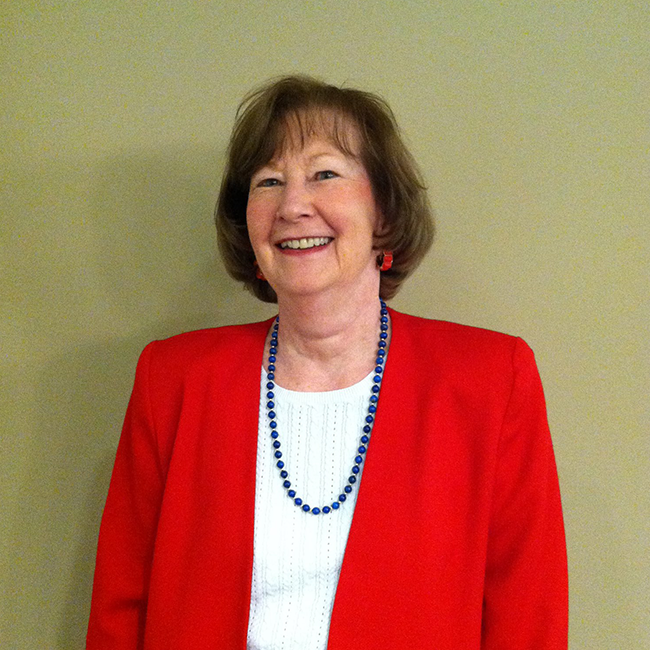 An image of an older adult woman with white skin and short bobbed brown hair. She is standing against a beige wall and smiling at the camera, wearing a red blazer, a white shirt, and a blue necklace.