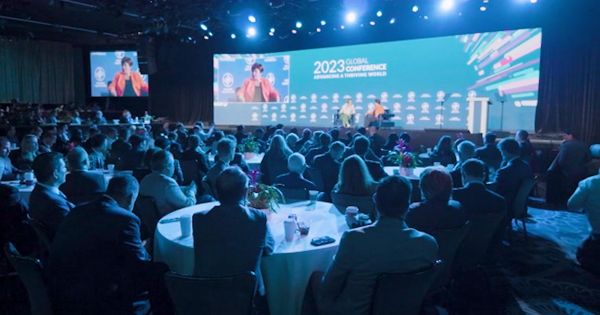 a dimly lit conference room full of attendees sitting at tables listening to a public speaker