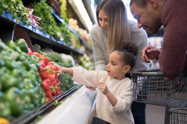 A male and female stand with young daughter in a grocery store aisle as she reaches for a red pepper in the produce section