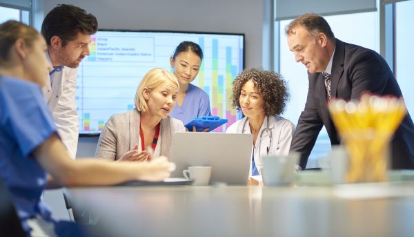 Six individuals sit around a conference table discussing something on a laptop screen in front of them