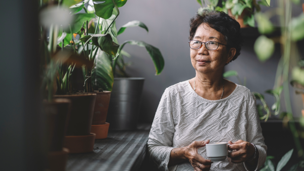 person sitting in room with plants, holding a mug of a beverage staring to their right