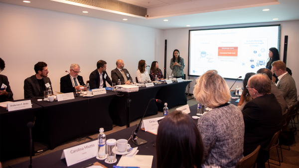 An image of a room full of business people and one presenter, all looking at an ambiguous business slide on a screen.