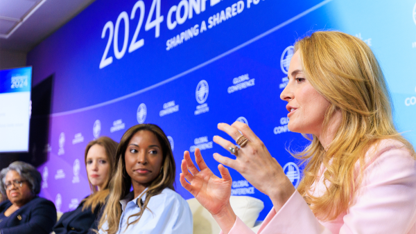 An image of four women sitting down and talking, on a stage at the Milken Institute's 2024 Global Conference. The women closest to the camera who is actively speaking has her hands raised, and she has pale white skin and long blonde hair. She is wearing a pink blazer. The other women are facing her as she speaks. 