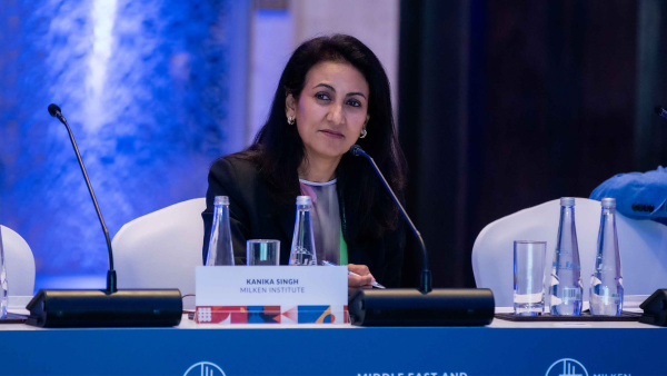 An image of an adult woman with tan skin and dark hair, wearing business attire and sitting in front of a microphone on a panel for a Milken Institute event.