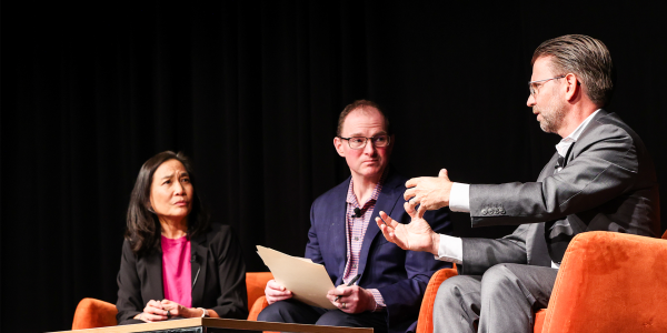 An image of three adults speaking on a stage, sitting down in orange chairs and wearing business attire. There are two men and one woman, and they are discussing the Future of Aging and Health.