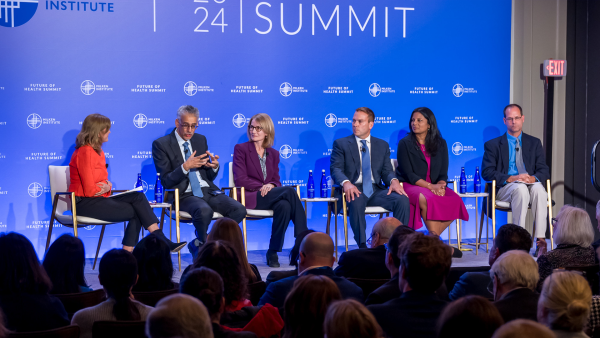 An image of six adults sitting in front of an audience on a stage, having a discussion. The backdrop is the Milken Institute's "2024 Future of Health Summit", and the adults are wearing business attire.