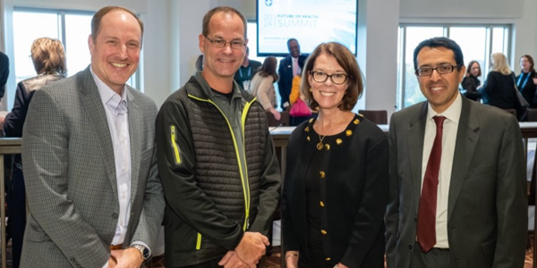 An image of four adults at a Future of Health and Aging conference, smiling and standing for a picture. There are 3 men and 1 woman and they are all wearing business attire. They are the Future of Aging Board Members for the Milken Institute.