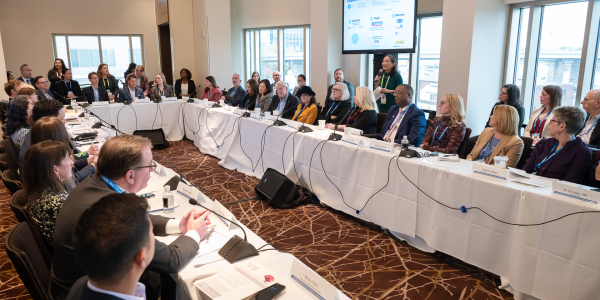 An image of several business people sitting at a panel at white tables in the shape of a rectangle, discussing the Future of Aging and Health. 