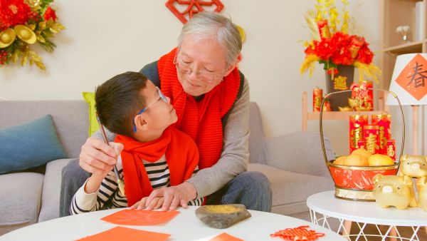 An image of a grandmother and young grandchild who are Asian in appearance, sitting at the kitchen table. The grandmother is hugging him and they are looking at each other. The house appears to be decorated for Lunar New Year, and they are wearing red scarves. This image leads to a newsletter about the Future of Aging by the Milken Institute.