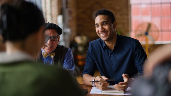 three people of diverse ages, gender, and ethnicities sitting at a table talking