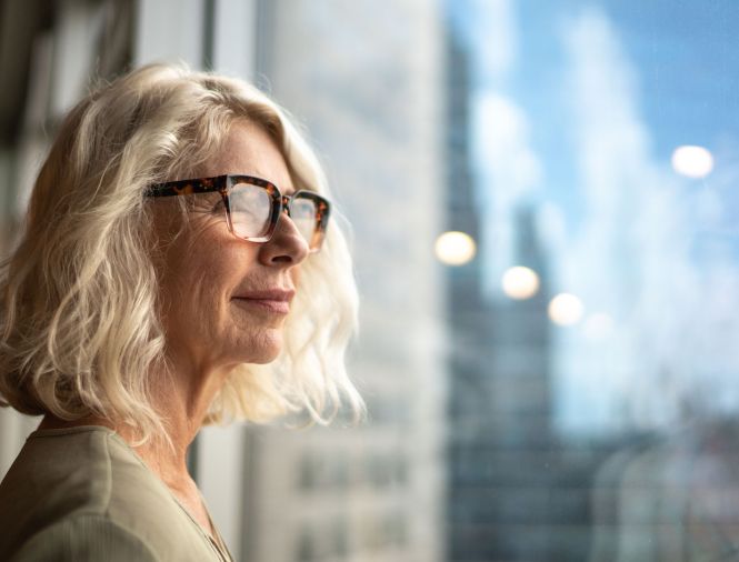 An older woman with blonde hair looks out a window of a highrise