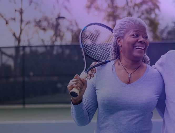 An older man and woman laugh with arms around each other while playing tennis