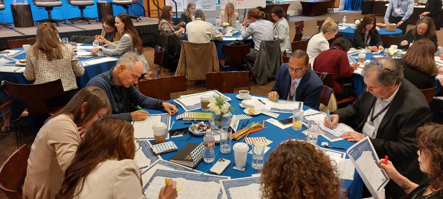 group of people sitting at a table with heads down writing a Milken Institute conference