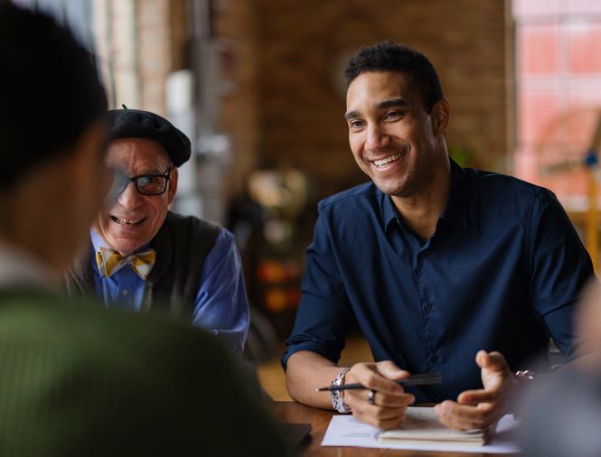 three people of diverse ages, gender, and ethnicities sitting at a table talking