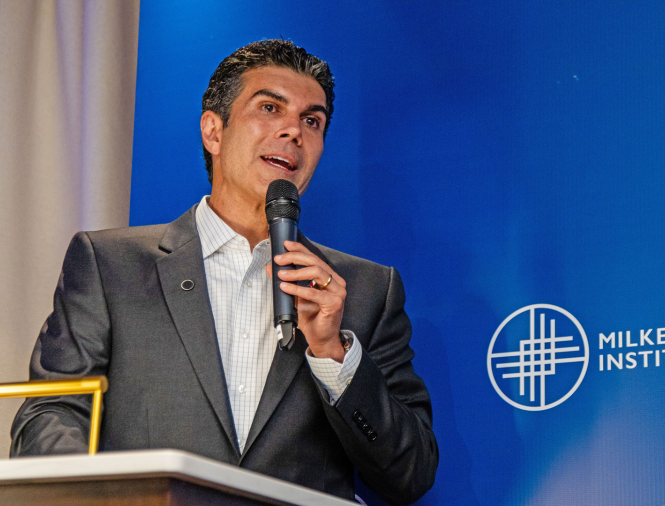 And image of a man with tan white skin and black/gray hair, holding a microphone and speaking on a stage, wearing a gray suit. He is speaking at the Milken Institute's Global Dinner Dialogues in Brazil and is standing in front of a blue "Milken Institute" background.