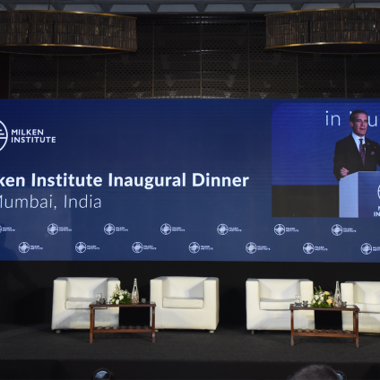 An image from the Milken Institute's Inaugural Dinner in Mumbai, India. It is an image of a speaker on a stage who is an adult man with white skin wearing a suit. There is a blue backdrop that reads "Milken Institute Inaugural Dinner" on the stage.