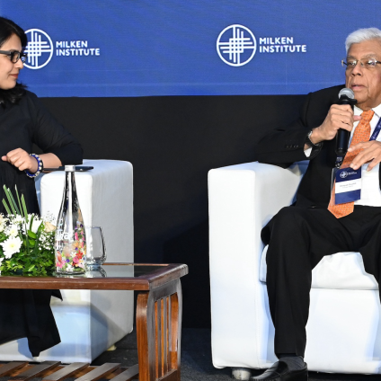 An image from the Milken Institute's Inaugural Dinner in Mumbai, India. There are two speakers sitting on a stage, one older man with white hair holding a microphone and speaking and one adult woman with dark hair watching him speak. Both have tan skin, are wearing business attire, and are Indian in appearance. There is a blue backdrop on the stage that reads "Milken Institute" with the company's logo, and the speakers are sitting in white chairs.