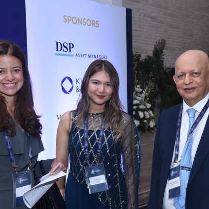 An image from the Milken Institute's Inaugural Dinner in Mumbai, India. There are two adult women with tan skin and dark hair standing next to a man with tan skin, and they are potentially Indian in appearance. All three people are wearing business attire and standing in front of event promotions that read "Milken Institute" followed by event information.