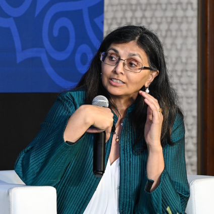 An image from the Milken Institute's Inaugural Dinner in Mumbai, India. A woman who is Indian in appearance with dark hair, tan skin, glasses, and a teal jacket is speaking and holding a microphone.