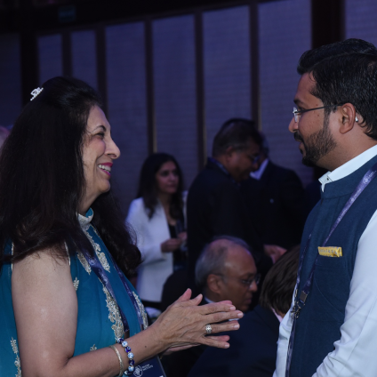 An image from the Milken Institute's Inaugural Dinner in Mumbai, India. Two adult people are talking to each other, one is a woman with tan skin and dark hair wearing a blue shirt and the other is a man with tan skin, dark hair, and glasses wearing a suit. They are both Indian in appearance and there are people in the background of what seems to be some sort of mixer or event.