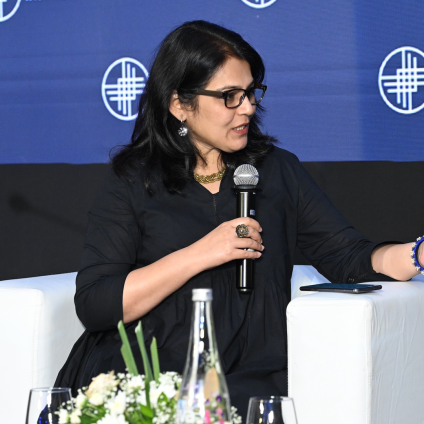 An image from the Milken Institute's Inaugural Dinner in Mumbai, India. There is an adult woman speaking on the stage, sitting in a white chair, and holding a microphone. She is Indian in appearance and has dark hair, tan skin, and a dark blue blazer. The backdrop is blue and has the Milken Institute's name and logo.