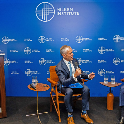 An image from the Milken Institute's Global Dinner Dialogues hosted in Brazil. The Milken Institute's CEO, Richard Ditizio is speaking with someone on a stage with a blue "Milken Institute" themed background and sitting near a podium. The image shows Richard Ditizio and another person, two adult men who have gray hair, white skin, and are wearing blue suits.