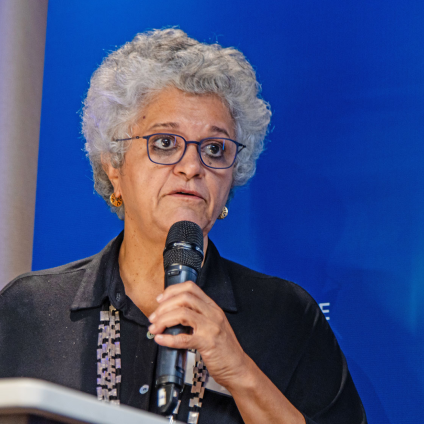 An image of an older adult woman with short curly gray and white hair, tan white skin, and glasses, holding a microphone and wearing a black shirt. She is speaking at the Milken Institute's Global Dinner Dialogues in Brazil and has a blue Milken Institute themed backdrop behind her.