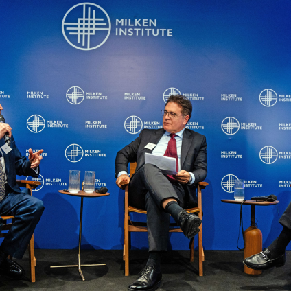 An image of three men sitting on a stage with a blue "Milken Institute" backdrop, speaking at the Milken Institute's Global Dinner Dialogues in Brazil. Each man is wearing a blue or grey-ish suit with a tie. The man on the furthest left has white hair and white skin, and is holding a microphone and speaking. The man in the middle has brown hair and white skin, and the man on the other end of the stage has white hair and white skin.