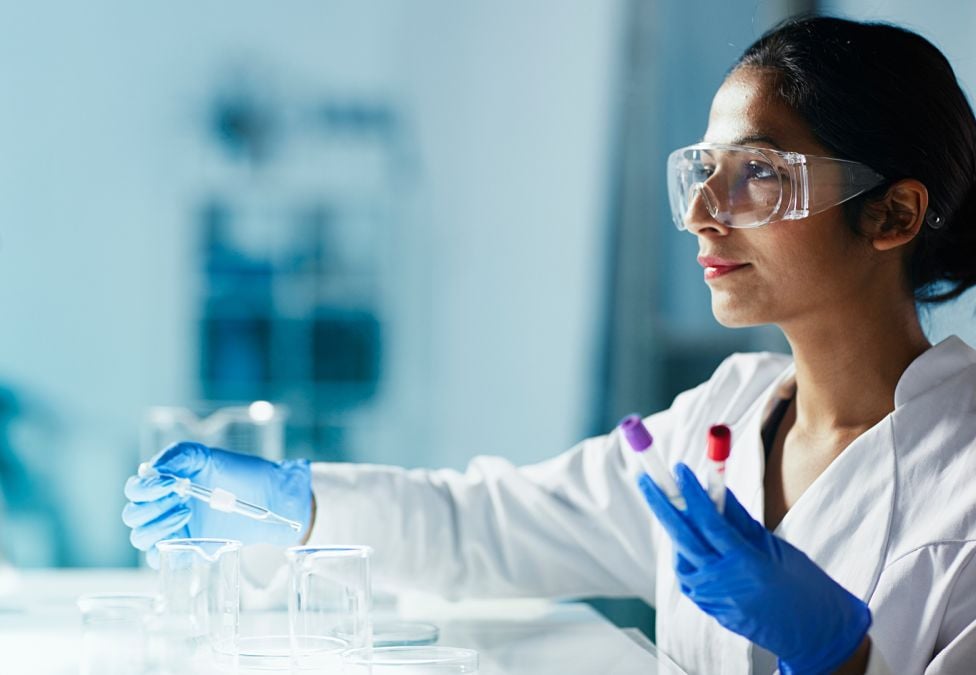 A female lab scientist with dark brown hair wearing safety goggles working with test tubes