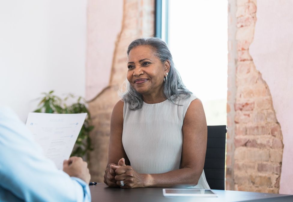 A smiling, darker skinned female with gray hair wearing a cream dress interviews at a table