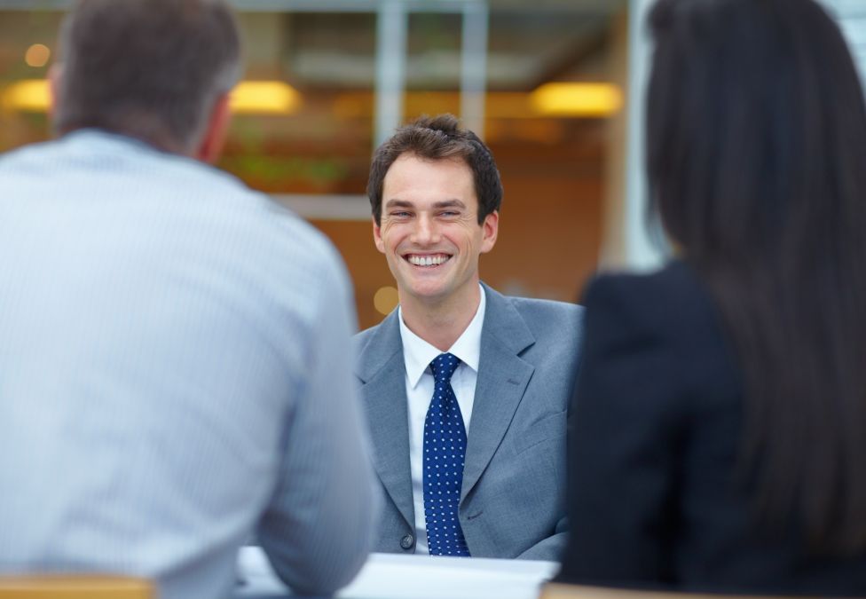 A smiling light skinned male in a suit and tie interviews with two other individuals