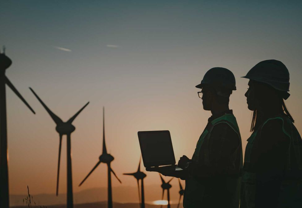 two workers wearing hard hats looking and windmills 