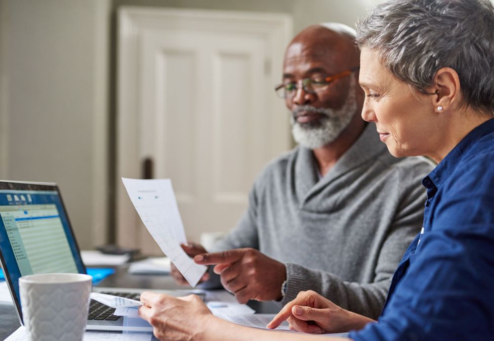 An elderly couple read paperwork in front of a laptop.